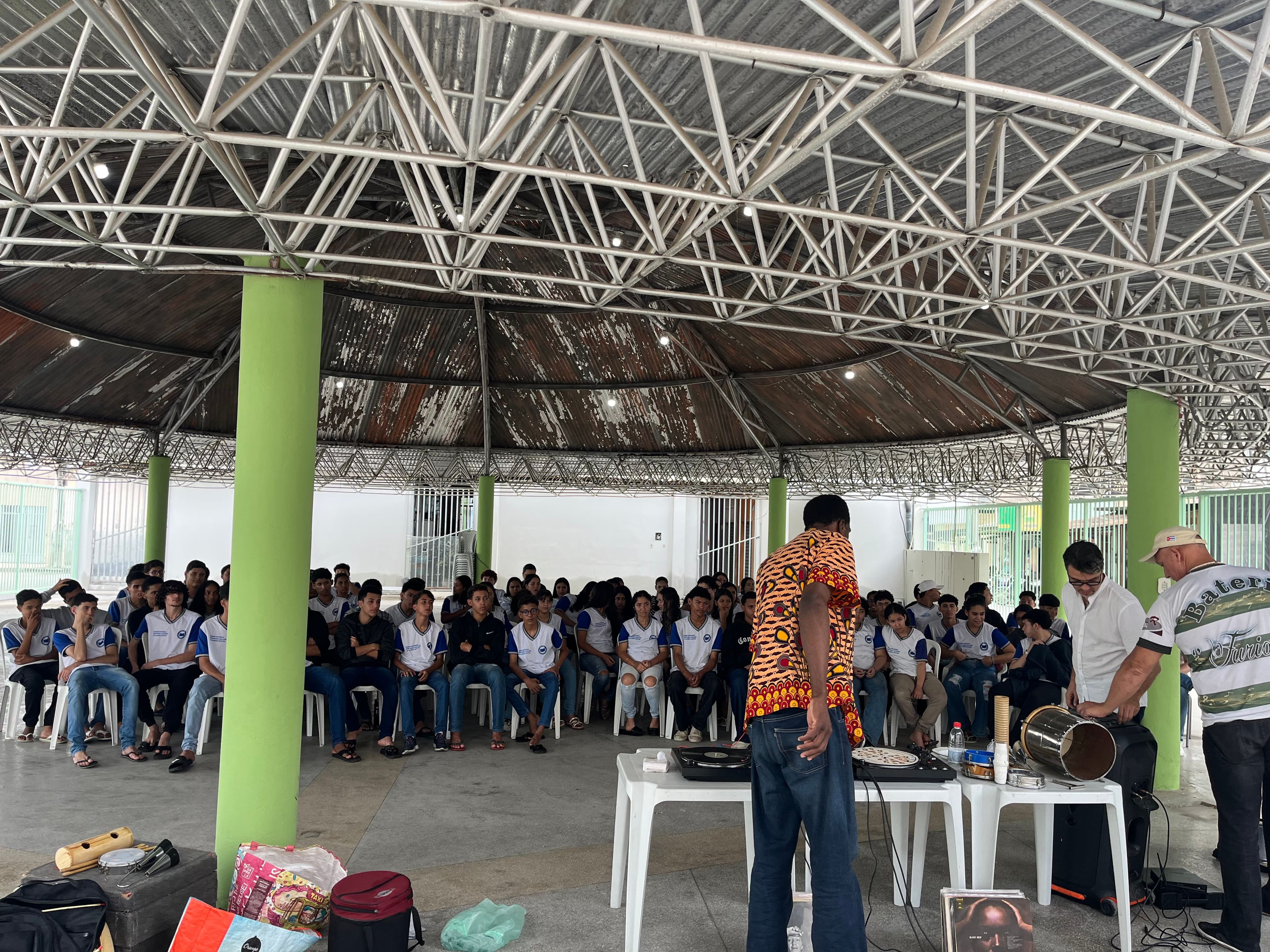 Encontro de potências: Tiganá Macedo no comando dos toca-discos para o "Dialogando ao Som do Vinil", enquanto a equipe prepara os instrumentos para a oficina de percussão.